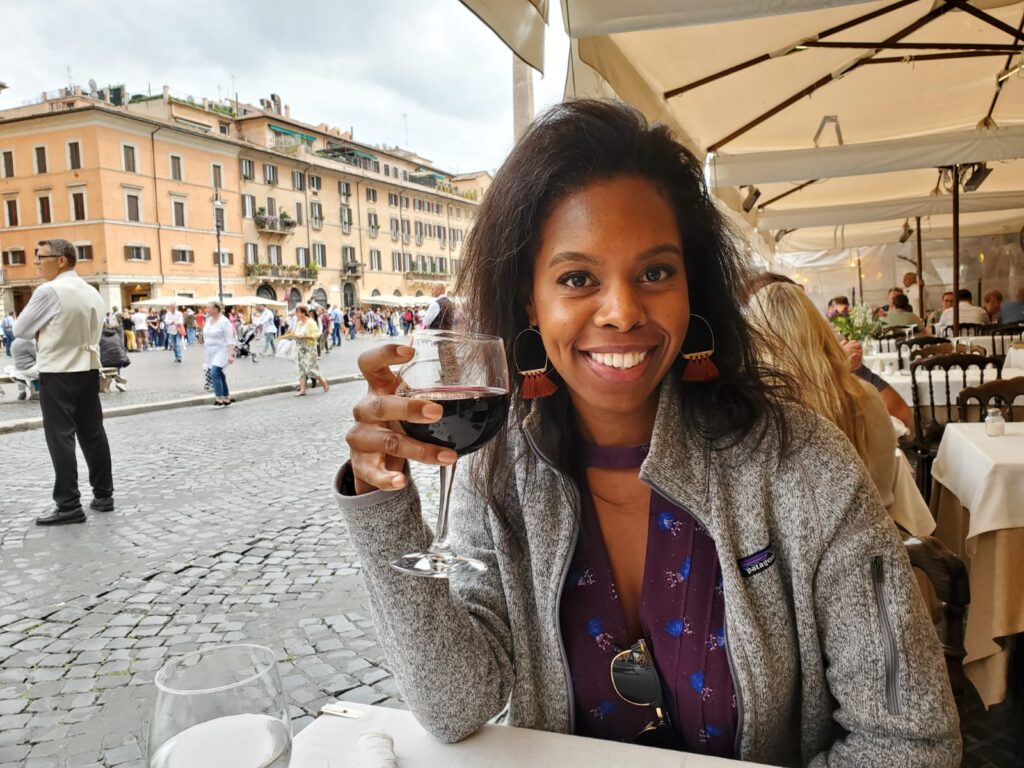 A beautiful black woman seated at Italian restaurant smiling and holding a glass of red wine. She is wearing a purple dress from Free People and a gray Patagonia full zip.