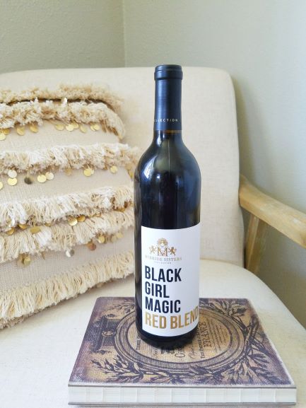A bottle of McBride Sisters Black Girl Magic wine sits on a French journal on a bohemian arm chair with fringe pillow in the background as decoration.